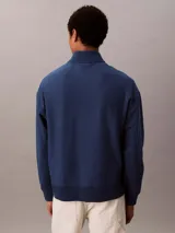 Buzo Calvin Klein de color azul marino, confeccionado en mezcla de algodón y poliéster. Cuenta con cuello alto, cierre de media cremallera y manga larga con hombros caídos. Presenta puños y dobladillo acanalados, y el logo de la marca bordado en punto de cadeneta en el pecho.