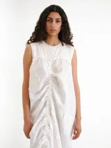 Vestido midi blanco sin mangas, con cuello redondo y diseño holgado. Presenta un cordón ajustable fruncido en el centro de la parte frontal y parches circulares sutiles en la tela.