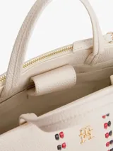 Cartera tipo satchel de Tommy Hilfiger en color beige, confeccionada en poliuretano con textura granulada. Presenta doble asa superior, correa ajustable extraíble, logo TH metalizado en el frente y dos franjas verticales decorativas con detalles entrelazados en rojo y negro.