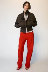 Cazadora tipo bomber de tejido efecto ante color marrón oscuro, con cuello subido, manga larga y cierre frontal con cremallera y botones ocultos. Presenta bolsillos frontales con solapa y botones metálicos.