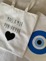 Tote bag de tela color blanco con estampado frontal de texto y corazón negro. Cuenta con cierre principal y bolsillo interno.