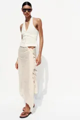 Pañuelo o foulard de tejido ligero color crudo o beige claro, con finas líneas verticales tejidas en un tono más claro. El borde inferior está decorado con borlas o pompones en tonos contrastantes como fucsia, morado y beige/dorado.