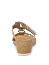 Sandalias de plataforma baja con diseño de tiras cruzadas en color beige y dorado. Presentan una tira superior con cierre de velcro y una tira frontal con detalle de calados geométricos y pequeñas tachas metálicas. La plantilla es acolchada y la suela tiene textura tipo yute.