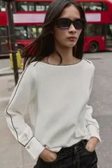 Blusa de crepe con cuello barco y manga larga. Presenta un diseño minimalista en color blanco con detalles de vivos en color negro que recorren el cuello, las mangas y los puños. Los puños incluyen botones decorativos.