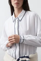 Camisa blanca de manga larga con detalles bordados en contraste a lo largo de la botonadura, el cuello y los puños. Presenta una silueta relajada y un diseño clásico con un toque artesanal.