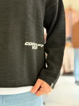 Sweater de punto liviano color beige, con textura marcada, cuello redondo y mangas largas de corte relajado, con pequeño estampado tipográfico negro en el lateral inferior.