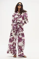 Pantalón ancho de lino con estampado floral grande en tonos morados sobre fondo blanco/crudo. Presenta cintura con elástico o ajuste y corte holgado en pierna.