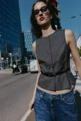 Chaleco jaspeado en tonos grises, con cuello redondo y sin mangas. Presenta cierre frontal con botones dorados y un cinturón fino negro con hebilla dorada.