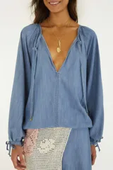 Blusa de mezclilla ligera con escote en V pronunciado, mangas largas amplias con puños ajustables mediante cordón y detalles de tiras colgantes en el cuello.