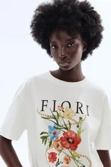 Remera oversized de algodón color blanco, con cuello redondo, mangas cortas y hombros caídos. Presenta un estampado frontal con la palabra 'Fiori', ilustraciones de flores coloridas y cadenas doradas.
