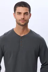 Remera de algodón texturada color gris oscuro, de manga larga, con cuello redondo y botones en la parte frontal. Presenta un pequeño logo bordado en el pecho y un parche decorativo en la manga izquierda.