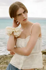 Top blanco de escote recto y tirantes finos ajustables, con detalle de lazos a contraste y flores bordadas. Cierre frontal con botones forrados en el mismo tejido.