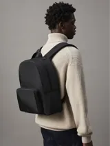 Mochila negra de poliéster reciclado con cierre de cremallera central, asa superior y correas de hombros acolchadas. Tiene un compartimento principal con cremallera, un bolsillo exterior en la parte delantera, funda para portátil y parte trasera de malla acolchada.
