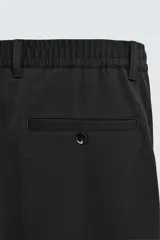 Pantalón de corte recto confeccionado en tejido elástico, con cintura elástica, pliegues frontales, bolsillos laterales y bolsillos traseros de vivo con botón.