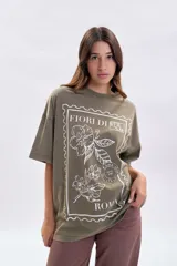 Remera de manga corta con corte oversize, color verde oliva con efecto de teñido especial. Presenta una estampa frontal en color blanco que simula un sello postal con motivos florales y la inscripción 'Fiori di Roma'.
