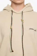 Sudadera larga de corte recto color beige, con escote redondo y capucha ajustable. Presenta mangas largas con forma, logo estampado en el pecho y cordones personalizados con nudos.