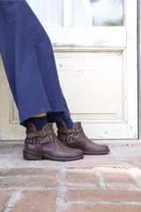 Bota corta de cuero color marrón, con taco bajo cuadrado y punta redondeada. Presenta un diseño estilo western con correas ajustables con hebillas metálicas y herrajes circulares en los laterales.