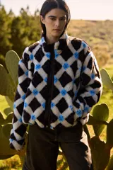 Campera de textura tipo corderito con diseño a cuadros en tonos marrones, negros y beige. Presenta cuello alto, cierre frontal completo y detalles en tela técnica lisa en los laterales y bordes. Posee un elástico ajustable en la parte inferior para regular el calce.