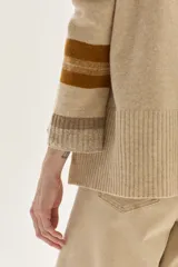 Sweater tejido de lambswool color beige, con cuello redondo y puños acampanados con rayas de colores (marrón, mostaza, bordó y beige). Incluye un detalle de temporada hecho a mano en la espalda.