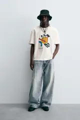 Camiseta de manga corta y cuello redondo, color blanco hueso, con un estampado abstracto multicolor en el pecho que incluye texto en la parte inferior.