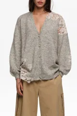 Cardigan gris melange de punto con escote en V y cierre frontal de botones. Presenta aplicaciones de encaje en tono rosado en el hombro izquierdo y en el lateral inferior derecho.