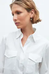 Camisa blanca de manga larga con cuello clásico y cierre frontal con botones. Presenta dos bolsillos plaqué en el pecho.