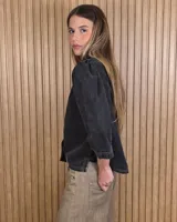 Camisa de denim color negro con efecto gastado, cuello alto con volados sutiles, mangas tres cuartos abullonadas en los hombros y cierre frontal con botones metálicos y costuras pespunteadas en contraste.