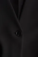 Blazer de punto grueso negro, con escote en V profundo sin cuello, hombreras y cintura entallada. Presenta cierre de un botón frontal y bolsillos delanteros con solapa.