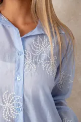 Camisa de manga larga en color blanco, confeccionada en tejido traslúcido con delicados bordados florales en hilo de seda al tono. Presenta cuello clásico, cierre frontal con botones y puños abotonados.