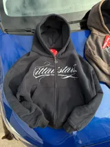 Campera con capucha negra, con cierre frontal completo y bolsillos estilo canguro. Presenta un estampado gráfico en blanco con letras cursivas en el pecho y diseño holgado.