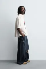 Camisa de algodón con estructura canvas, color rojo lavado, de corte relajado (relaxed fit) y manga corta. Posee cuello solapa, dos bolsillos de plastrón en el pecho y cierre frontal de botonadura. Presenta bordado y estampado de texto a contraste en el delantero.