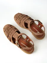 Sandalias estilo cangrejera color marrón, confeccionadas en cuero con diseño trenzado en la parte delantera, tira ajustable con hebilla metálica en el tobillo y suela plana.