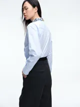 Camisa de algodón color celeste claro, con corte clásico, cuello camisero y cierre frontal de botones. Presenta apliques decorativos de pedrería y piezas metálicas circulares en la parte frontal.