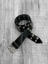 Cinto negro de charol con hebilla metálica plateada y ojales metálicos plateados.