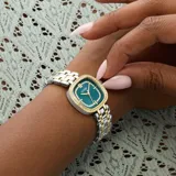 Reloj analógico Guess modelo Hazel con caja cuadrada de 25mm y correa tipo brazalete de acero inoxidable en tono combinado plateado y dorado. La esfera color verde teal presenta un acabado sunray, manecillas doradas y un marco decorado con cristales brillantes.