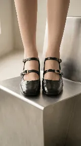 Zapatos tipo Mary Jane de cuero negro con acabado brillante, punta redondeada, taco bajo cuadrado y doble tira con hebillas metálicas doradas en el empeine.