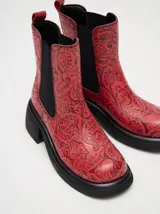 Bota tipo chelsea de cuero vacuno en color rojo con estampado floral grabado en negro. Presenta paneles laterales elásticos, suela de plataforma y taco cuadrado en color negro.