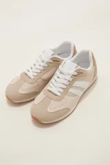 Zapatilla deportiva de estilo retro en tonos beige y crudo, con paneles de gamuza sintética y tela, cordones blancos y suela de goma color caramelo.