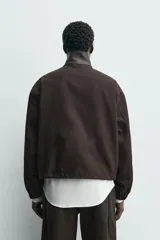 Cazadora de corte relajado en color marrón oscuro, confeccionada en tejido de algodón con cuello subido de piel. Presenta cierre frontal con cremallera, bolsillos laterales de vivo y mangas largas con puños elásticos.