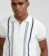 Camisa polo slim de punto, color blanco con rayas verticales oscuras en contraste en el frente y líneas en los bordes de las mangas cortas. Cuenta con cuello polo y cierre frontal parcial con cremallera.