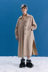 Trench midi de color camel, confeccionado en una mezcla de algodón y nylon con propiedades repelentes al agua. Presenta un diseño clásico con cuello camisero, cierre frontal de botones, bolsillos laterales y tajos en los costados.