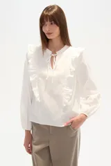 Blusa blanca de algodón con volados frontales, cuello redondo con lazo ajustable y mangas largas abullonadas con puños elásticos.