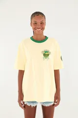 Camiseta Ipanema de manga corta en color amarillo pastel, con cuello redondo en contraste de color verde oscuro. Presenta un pequeño estampado en el pecho con diseño de escudo y palmeras.