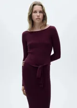 Vestido midi de punto acanalado en color bordó, con cuello barco, manga larga y diseño entallado. Incluye un cinturón desmontable de la misma tela para ajustar a la cintura y presenta una abertura lateral.