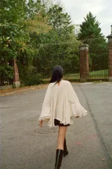 Top tipo capa de color beige, con cuello subido y cierre posterior con botones ocultos. Presenta un diseño fluido con bajo asimétrico y mangas acampanadas.