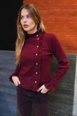Remera de manga larga color blanco, confeccionada en tejido liviano con caída fluida. Presenta cuello alto con volados, abotonadura frontal decorativa y detalles de volados a lo largo de la abertura, además de terminaciones con volados en los puños.