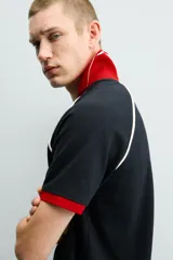 Polo de corte relajado confeccionado en tejido de algodón con estructura abierta. Presenta cuello polo con abertura frontal en V, mangas cortas tipo ranglán y acabados en rib a contraste en color rojo y blanco.