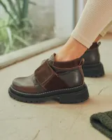 Botinetas de caña baja con suela track gruesa, combinando cuero negro en la puntera, gamuza marrón en el lateral y detalles de textura animal print tipo serpiente en tonos marrones. Presenta un cierre de solapa con velcro en el empeine.