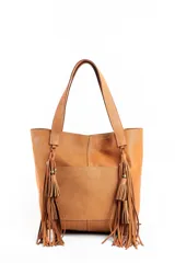 Cartera tipo tote bag color tostado, confeccionada en cuero perforado. Presenta doble asa de hombro y dos grandes borlas decorativas con flecos largos en los laterales. Incluye un bolsillo frontal externo y costuras visibles a contratono.