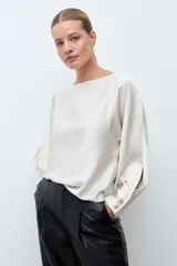 Blusa de satén color crudo, con cuello bote, mangas largas con aberturas laterales y puños abotonados.